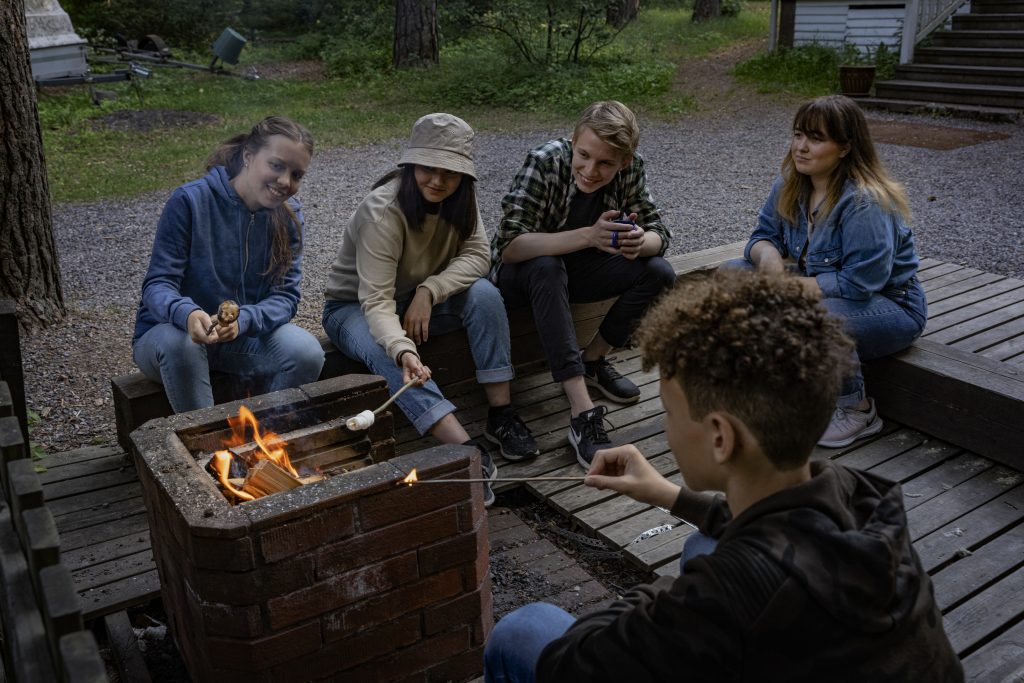 Viisi nuorta paistaa vaahtokarkkeja iltanuotiolla.