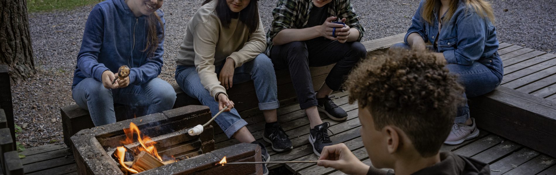 Viisi nuorta paistaa vaahtokarkkeja iltanuotiolla.