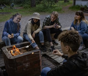 Viisi nuorta paistaa vaahtokarkkeja iltanuotiolla.