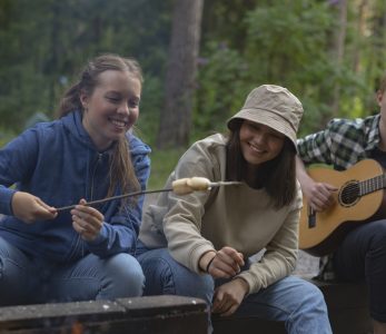 Nuoria nuotiolla, yksi paistaa vaahtokarkki, yksi nauraa ja yksi soittaa kitaraa.