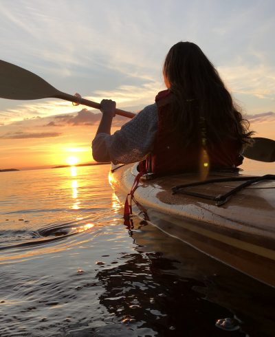 Nainen meloo kajakilla kohti auringonlaskua.