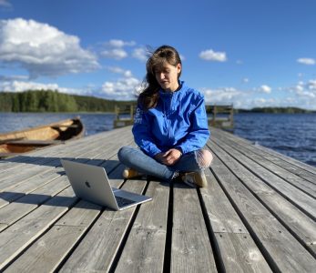 Nainen istuu laiturilla läppärin kanssa, aurinko paistaa.