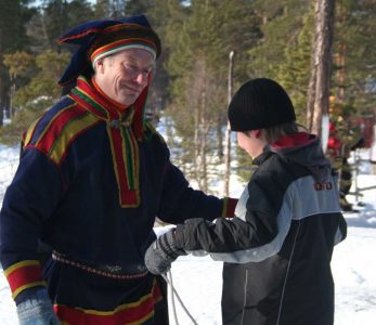 Saamelaiseen perinneasuun pukeutunut mies jututtaa nuorta hiihtäjää