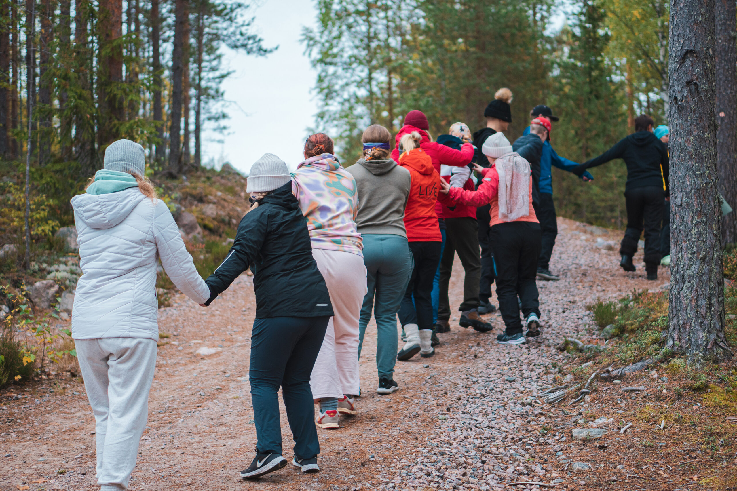 Nuoria jonossa pitämässä toisiaan käsistä kiinni. Jono liikkuu ylämäkeen soratietä pitkin.