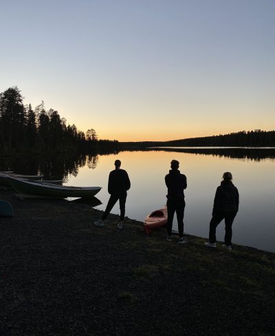 Silhuettikuvassa ihmisiä ja veneitä rannalla ilta-auringossa.