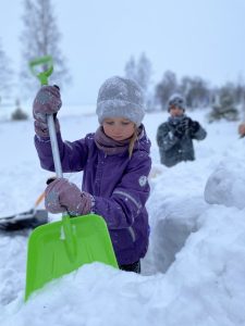 Lapsi lapioimassa lunta lumilinnaa varten. Taustalla toinen lapsi touhuissa mukana.
