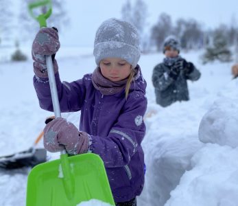 Lapsi lapioimassa lunta lumilinnaa varten. Taustalla toinen lapsi touhuissa mukana.