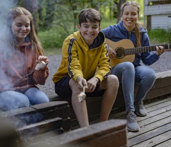 Kolme nuorta rippileirillä iltanuotiolla. Kaksi paistaa vaahtokarkkeja tikuilla, kolmas soittaa kitaraa.