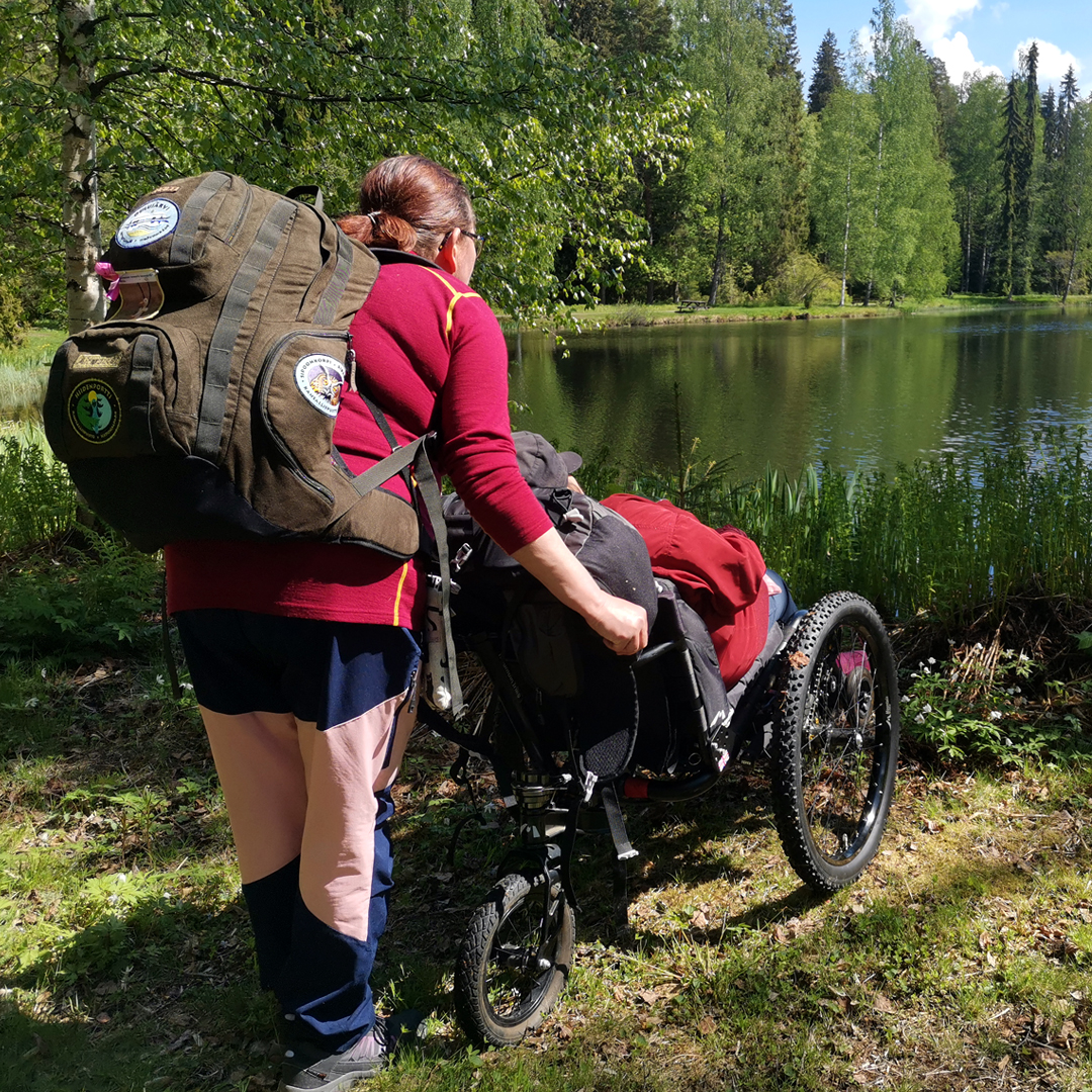Pyörätuolissa istuva saattajan kanssa järven rannalla. Pyörätuolissa on maastorenkaat.